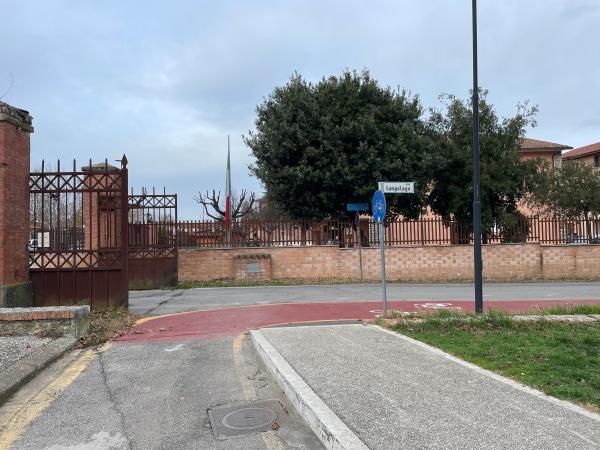 Eingang mit schmiedeeisernem Tor und roten Backsteinmauern. Roter Fahrradweg kreuzt das Bild